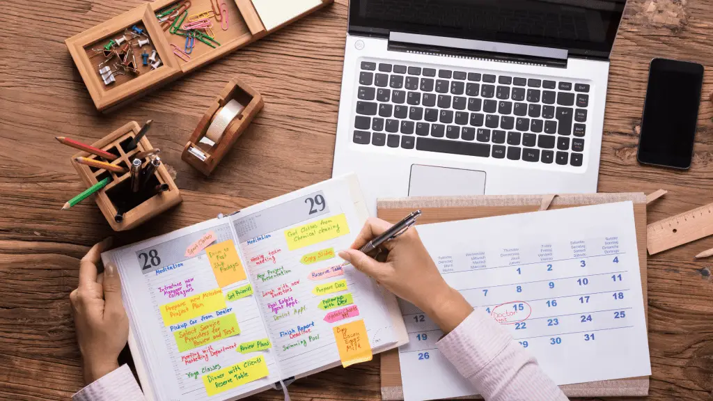 A desktop with a computer, office supplies, and iPhone, a calendar, and a planner with post it notes. A man is writing on a post it note in the planner, trying to figure out his back-to-school scheduling.