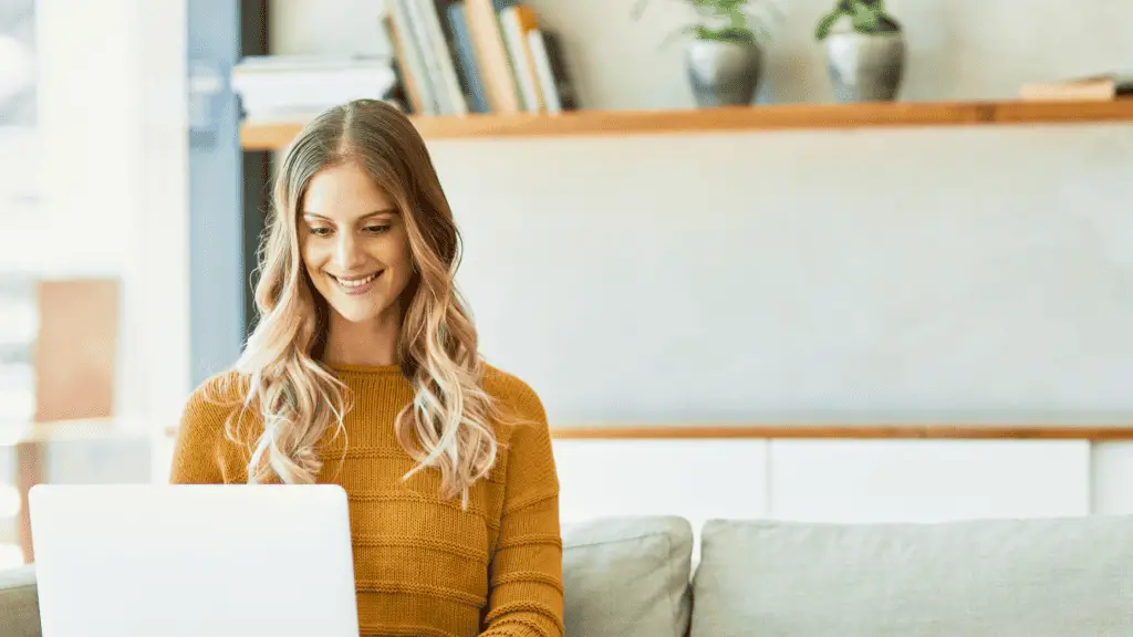 woman wearing a mustard colored sweater sitting on a white sofa typing a blog post on a white laptop.
