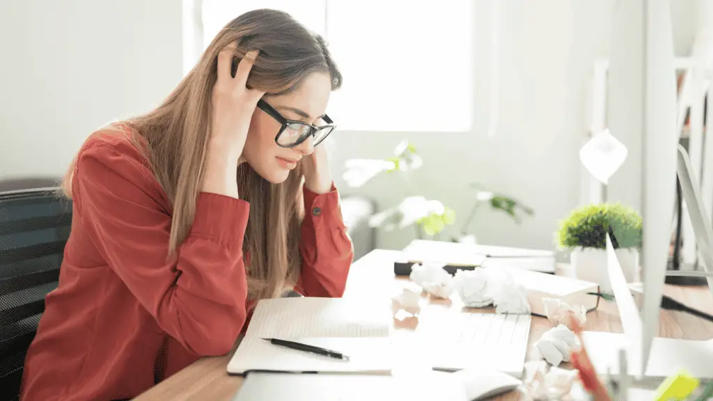 woman with writer's block is sitting at a desk with crumpled up paper, frustrated and in need of blog inspiration