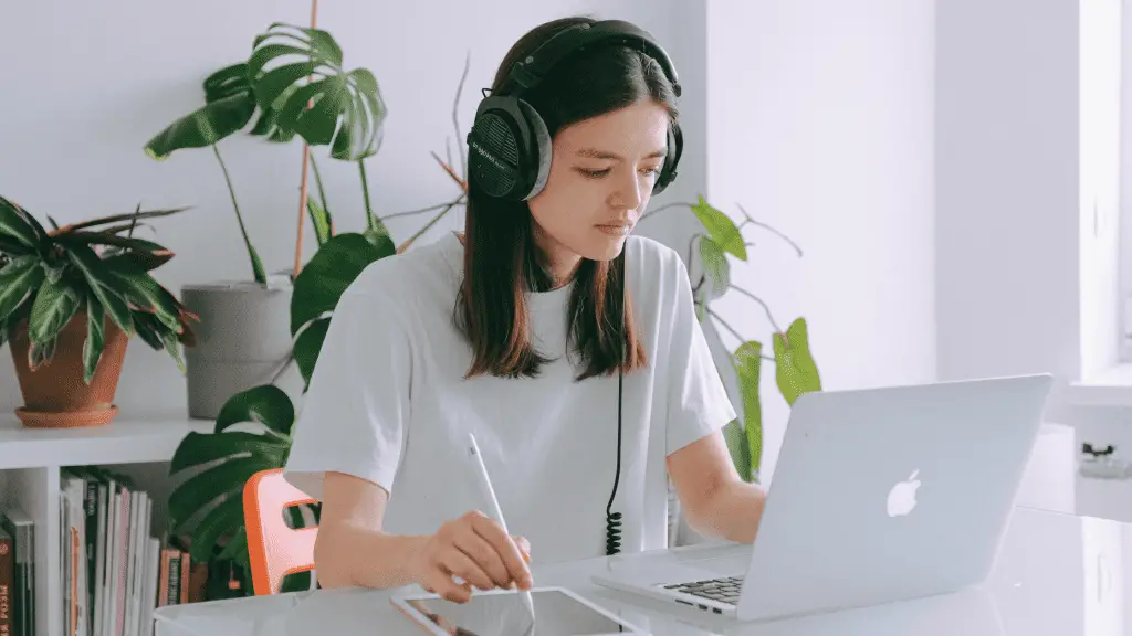 Woman freelancer wearing headphones, sitting at a desk working on her computer and drawing on an iPad.