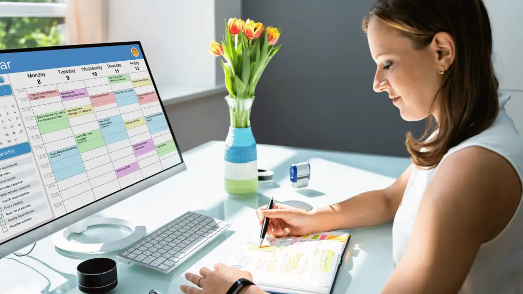 woman sitting at a desk looking at her content calendar on her computer and on a paper planner. There is a vase of orange tulips sitting to her right.